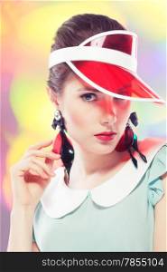 Young beautiful woman with red glamour lips and eye arrow make-up wearing fancy plastic earrings and red sun visor on multicolor background, retro beauty style