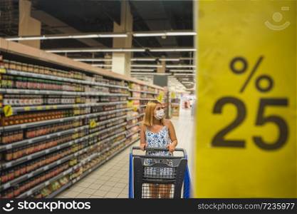 Young beautiful woman with shopping cart wears medical mask against coronavirus while grocery shopping and stockpiling in supermarket. Shopping concept during pandemic corona,covid19.. Young girl does shopping in supermarket