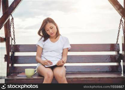 Young beautiful woman writing on notepad while sitting on the bench. Relaxing on the beach.