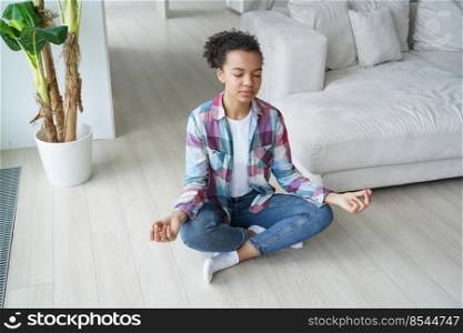 Young biracial girl doing yoga sitting in padmasana, lotus pose on floor indoors. African american woman meditating, doing breathing exercises at home Interior. Stress relief, wellness concept.. Young biracial girl practice yoga, meditate, do breathing exercises at home. Stress relief, wellness
