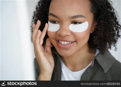Young biracial girl use under eye patches, close-up portrait. Smiling pretty mixed race teen lady moisturize face by collagen pads for healthy facial skin. Skincare treatment, beauty routine.. Young biracial girl applying under eye patches for healthy fresh face skin. Skincare, beauty routine