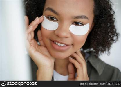 Young biracial girl with applied collagen patches on under eye skin smiling. Mixed race teen lady caring for under-eye skin, preventing puffiness, enjoying morning beauty routine. Skincare concept. Young biracial girl applying patches on under eye skin smiling. Morning beauty routine, skincare