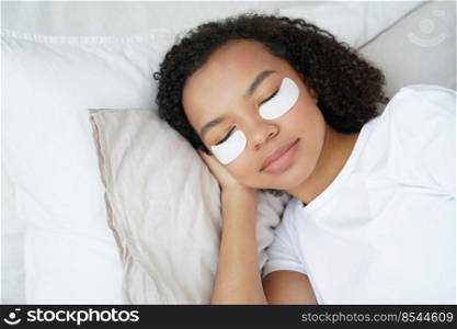 Young biracial girl with under eye patches on face relaxing lying in bed with closed eyes. Pretty woman using undereye skin beauty masks, preventing bags, reducing wrinkles. Morning skincare routine.. Young biracial girl with under eye patches on face relax lying in bed. Morning skincare routine