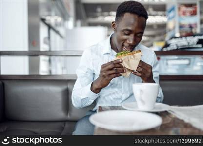 Young black businessman eating sandwich in car showroom. Successful business person on motor show, black man in formal wear on food-court. Black businessman eating sandwich in car showroom