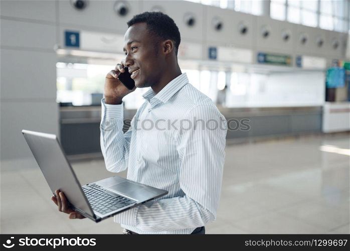 Young black businessman with laptop and phone negotiates in car showroom. Successful business person on motor show, black man in formal wear. Businessman with phone negotiates in car showroom