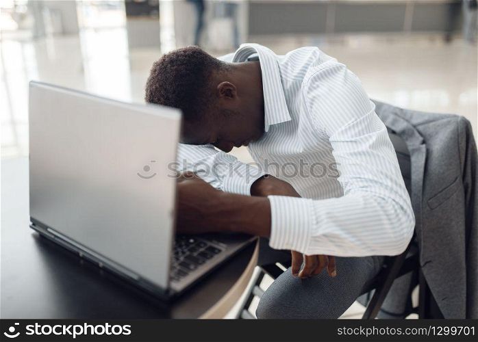 Young black businessman with laptop sleeping in office cafe. Tired business person drinks coffee in food-court, black man in formal wear. Businessman with laptop sleeping in office cafe