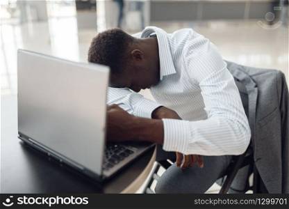 Young black businessman with laptop sleeping in office cafe. Tired business person drinks coffee in food-court, black man in formal wear. Businessman with laptop sleeping in office cafe
