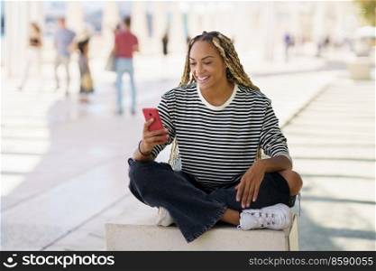 Young black female using a smartphone, wearing her hair in braids. Typical African hairstyle.. Black female using a smartphone sitting on a bench outdoors, wearing her hair in braids.