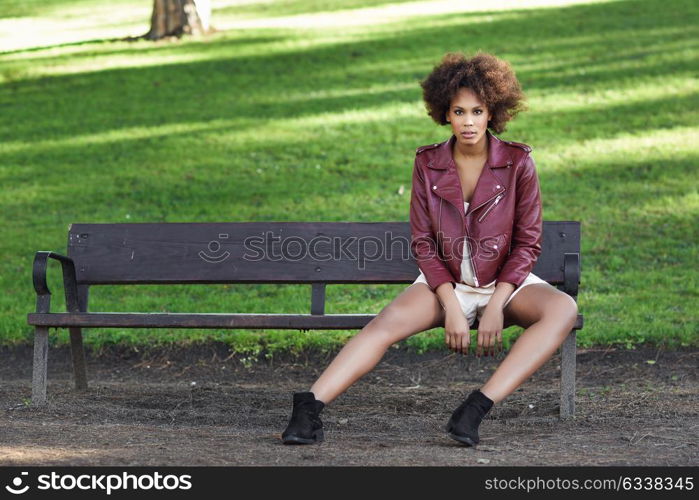 Young black female with afro hairstyle sitting in a bench in an urban park. Mixed woman wearing red leather jacket and white dress with city background.