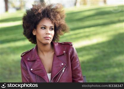 Young black female with afro hairstyle sitting in a bench in an urban park. Mixed woman wearing red leather jacket and white dress with city background.