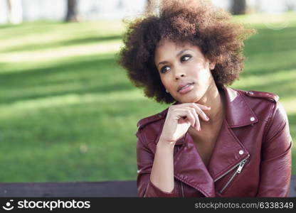 Young black female with afro hairstyle sitting in a bench in an urban park. Mixed woman wearing red leather jacket and white dress with city background.