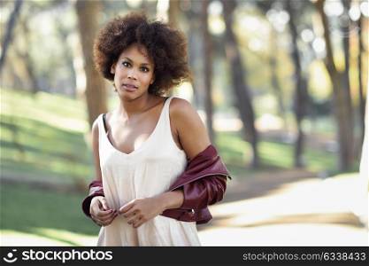 Young black female with afro hairstyle standing in an urban park. Mixed woman wearing red leather jacket and white dress with city background.