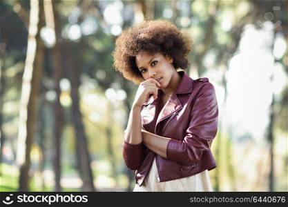 Young black female with afro hairstyle standing in an urban park. Mixed woman wearing red leather jacket and white dress with city background.