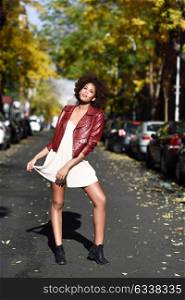 Young black female with afro hairstyle standing in an urban street. Mixed woman wearing red leather jacket and white dress with city background.