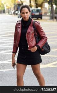 Young black female with afro hairstyle standing in an urban street. Mixed woman wearing red leather jacket, black sweater and skirt with city background. Female carrying bag and smartphone.
