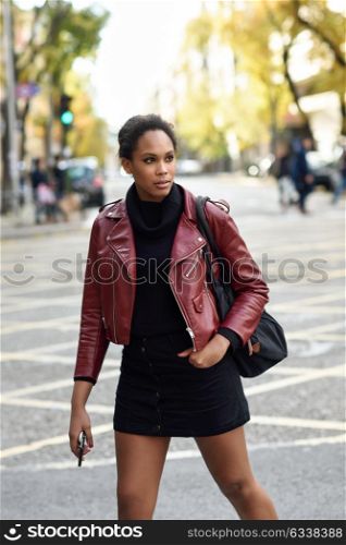 Young black female with afro hairstyle standing in an urban street. Mixed woman wearing red leather jacket, black sweater and skirt with city background. Female carrying bag and smartphone.