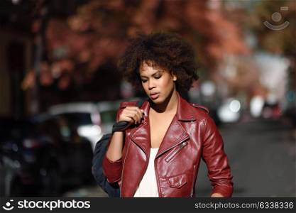 Young black female with afro hairstyle standing in an urban street carrying a bag. Mixed woman wearing red leather jacket and white dress with city background.