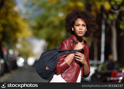 Young black female with afro hairstyle standing in an urban street carrying a bag. Mixed woman wearing red leather jacket and white dress with city background.