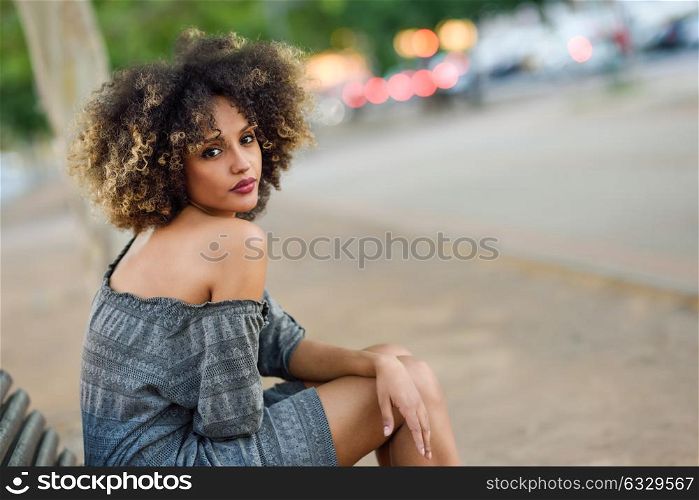 Young black girl with afro hairstyle sitting on a bench in urban background. Mixed woman wearing casual clothes