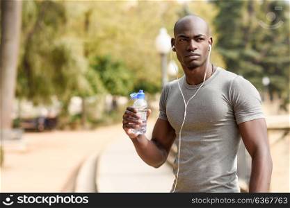 Young black man drinking water before running in urban background. Young male exercising with naked torso listening to music with headphones.