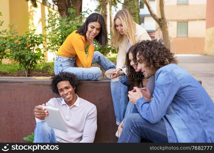 Young black man showing something on his digital tablet to his group of friends sitting on some steps in the street.. Multi-ethnic group of young people looking at a digital tablet outdoors in urban background.