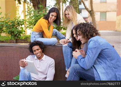 Young black man showing something on his digital tablet to his group of friends sitting on some steps in the street.. Multi-ethnic group of young people looking at a digital tablet outdoors in urban background.