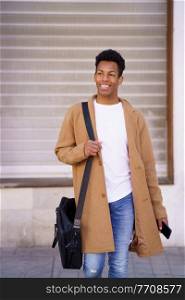 Young black man walking down the street carrying a briefcase and a smartphone in his hand. Cuban guy smiling in urban background.. Young black man walking down the street carrying a briefcase and a smartphone in his hand.