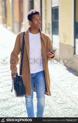 Young black man walking down the street carrying a briefcase and a smartphone. Cuban guy in urban background.. Young black man walking down the street carrying a briefcase and a smartphone.
