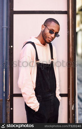 Young black man wearing casual clothes and sunglasses, against a pink urban background. African guy with bib pants standing outdoors. African guy with bib pants standing outdoors