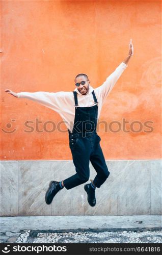 Young black man wearing casual clothes jumping in urban background. Lifestyle concept. Millennial african guy with bib pants outdoors