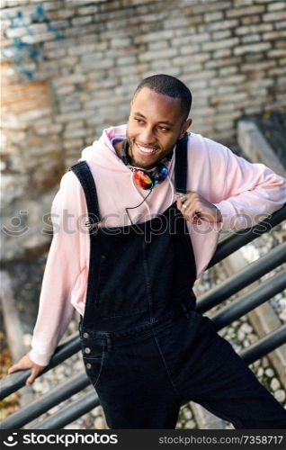 Young black man wearing casual clothes smiling on urban stairs. Happy African guy with bib pants outdoors. Smiling black man wearing casual clothes outdoors