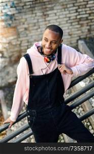 Young black man wearing casual clothes smiling on urban stairs. Happy African guy with bib pants outdoors. Smiling black man wearing casual clothes outdoors