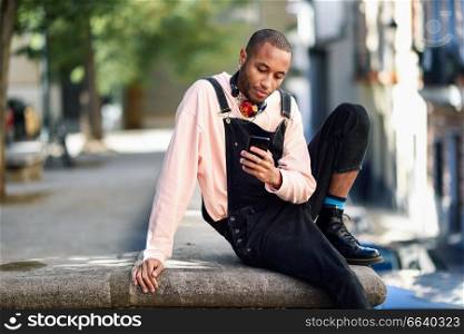 Young black man with headphones sitting in urban street looking at his smart phone. Lifestyle concept.