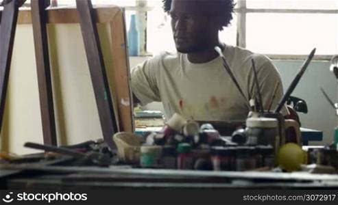 Young black people painting for hobbies. Proud african american man working as professional painter, skillful Cuban artist in art studio near table full of equipment, tools, canvas and paint