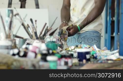 Young black people painting for hobby. African american man working as professional painter, skillful Cuban artist in art studio, cleaning brush and tools on table full of equipment, canvas and paint