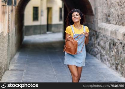 Young black tourist woman with curly hairstyle outdoors. Arab traveler girl in casual clothes in the street. Happy female wearing yellow t-shirt and denim dress in urban background.. Young black tourist woman with curly hairstyle outdoors
