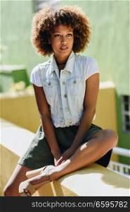 Young black woman, afro hairstyle, in the street. Girl, model of fashion, wearing casual clothes in urban background. Female with skirt and denim vest.