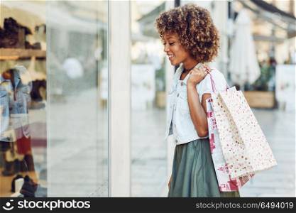 Young black woman, afro hairstyle, looking at a shop window. Young black woman in front of a shop window in a shopping street. African girl with afro hairstyle wearing casual clothes.