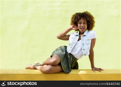 Young black woman, afro hairstyle, sitting on a wall smiling. Girl, model of fashion, wearing casual clothes in urban background. Female with skirt, denim vest and high heels.