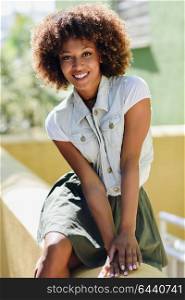 Young black woman, afro hairstyle, smiling. Girl, model of fashion, wearing casual clothes in urban background. Female with skirt and denim vest.