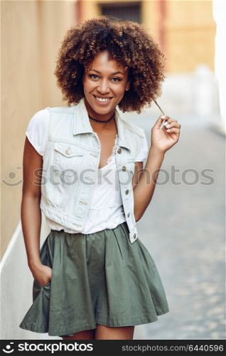 Young black woman, afro hairstyle, smiling in the street. Girl wearing casual clothes in urban background. Female with skirt and denim vest