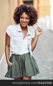 Young black woman, afro hairstyle, smiling in the street. Girl wearing casual clothes in urban background. Female with skirt and denim vest