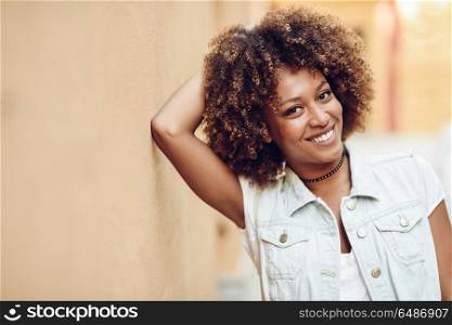 Young black woman, afro hairstyle, smiling in urban background. Young black woman, afro hairstyle, smiling near a wall in the street. Girl wearing casual clothes in urban background.