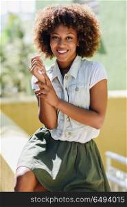 Young black woman, afro hairstyle, smiling in urban background. Young black woman, afro hairstyle, smiling. Girl, model of fashion, wearing casual clothes in urban background. Female with skirt and denim vest.