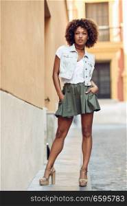 Young black woman, afro hairstyle, standing in the street. Girl wearing casual clothes in urban background. Female with skirt, denim vest and high heels.