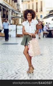 Young black woman, afro hairstyle, with shopping bags in the street. Girl wearing casual clothes in urban background. Female with skirt, denim vest and high heels.