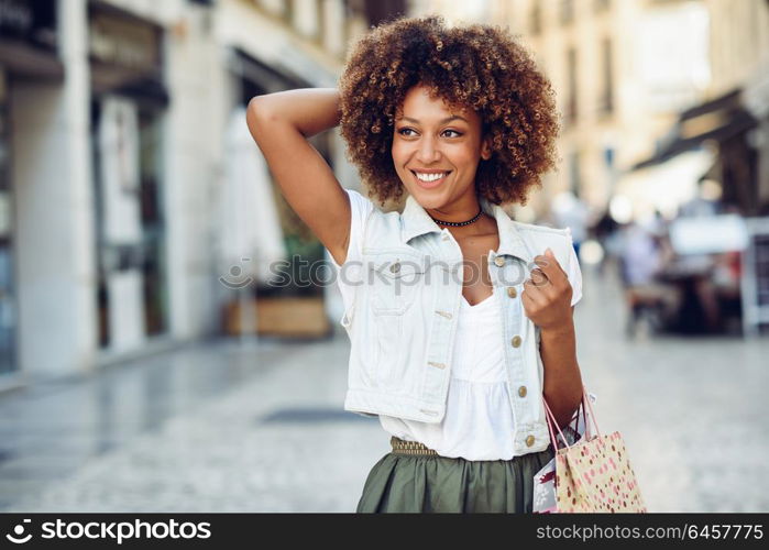 Young black woman, afro hairstyle, with shopping bags in the street. Girl wearing casual clothes in urban background. Female with skirt, denim vest and high heels.