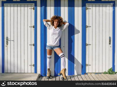 Young black woman on roller skates near a beach hut. Girl with afro hairstyle rollerblading on sunny promenade.