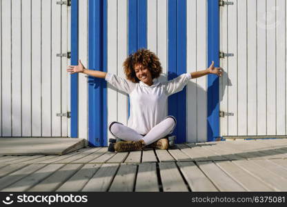 Young black woman on roller skates sitting near a beach hut. Girl with afro hairstyle rollerblading on sunny promenade.