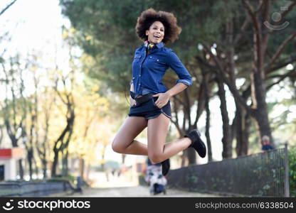 Young black woman with afro hairstyle jumping in urban background. Mixed woman wearing blue shirt and shorts. Female carrying funny headphones.